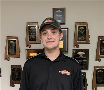 Photo of male employee wearing a SERVPRO hat and shirt in front of awards hanging on a wall